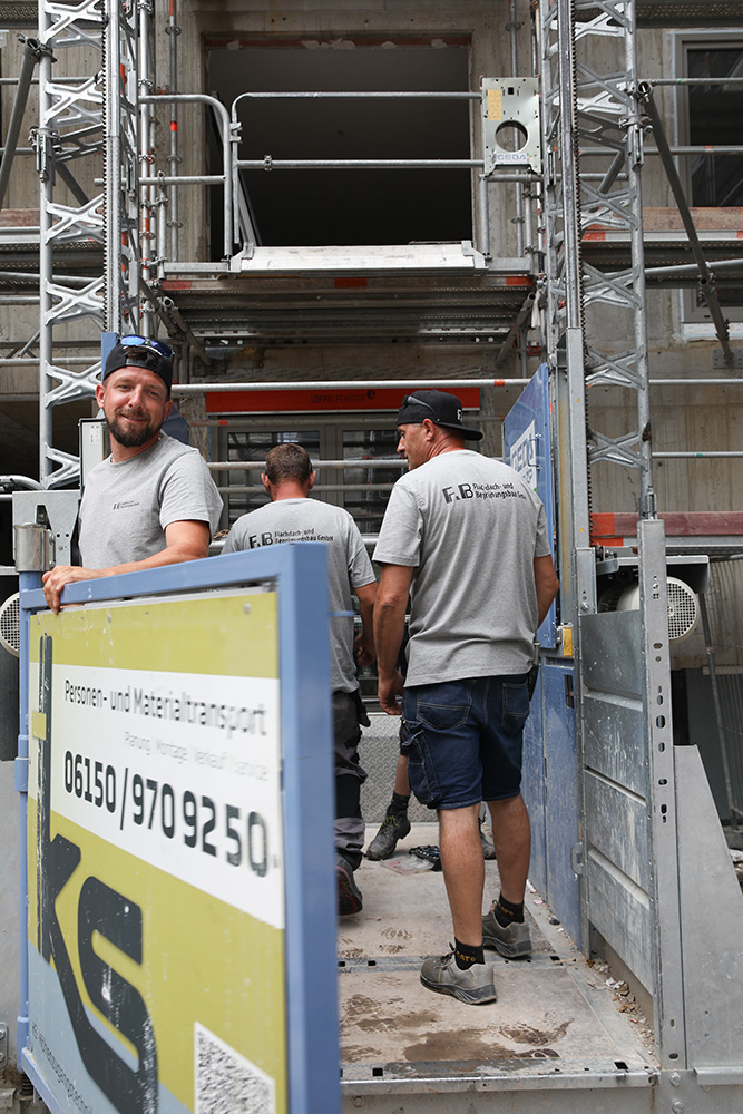 FuB-Dachdeckerteam an einem Baustellenaufzug vor einem eingerüsteten Gebäude